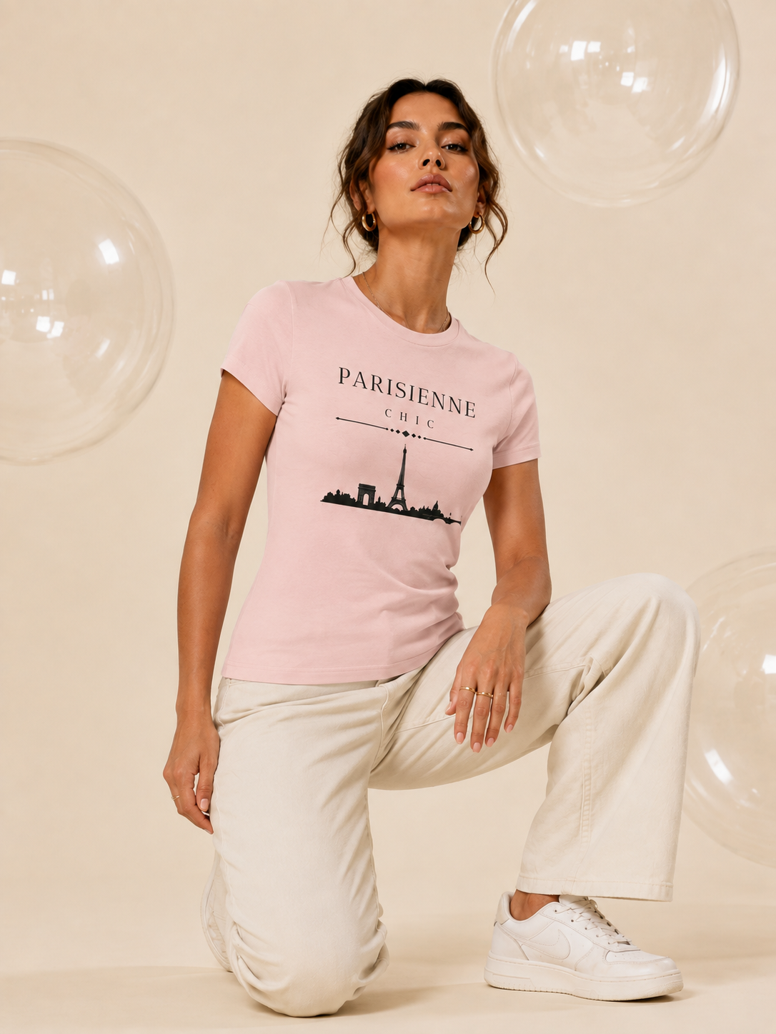 A woman kneeling on a beige studio floor wearing a light pink PARISIENNE CHIC graphic T-shirt with a black Paris skyline and Eiffel Tower print, paired with cream wide-leg trousers and white sneakers. Clear decorative bubbles float around her against a soft champagne-beige studio background.