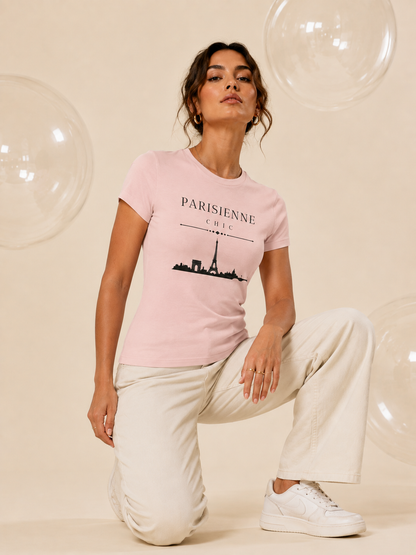 A woman kneeling on a beige studio floor wearing a light pink PARISIENNE CHIC graphic T-shirt with a black Paris skyline and Eiffel Tower print, paired with cream wide-leg trousers and white sneakers. Clear decorative bubbles float around her against a soft champagne-beige studio background.