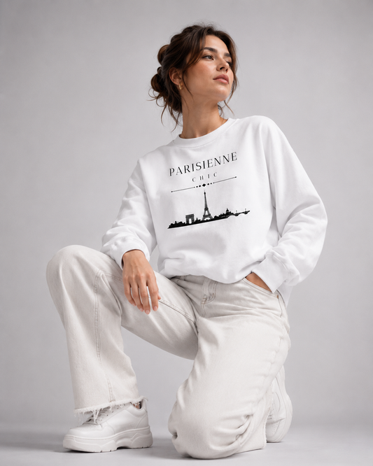 A woman kneels on a light gray studio floor wearing a white crewneck sweatshirt with a black 'PARISIENNE CHIC' and Eiffel Tower graphic, paired with off-white wide-leg jeans and white sneakers.