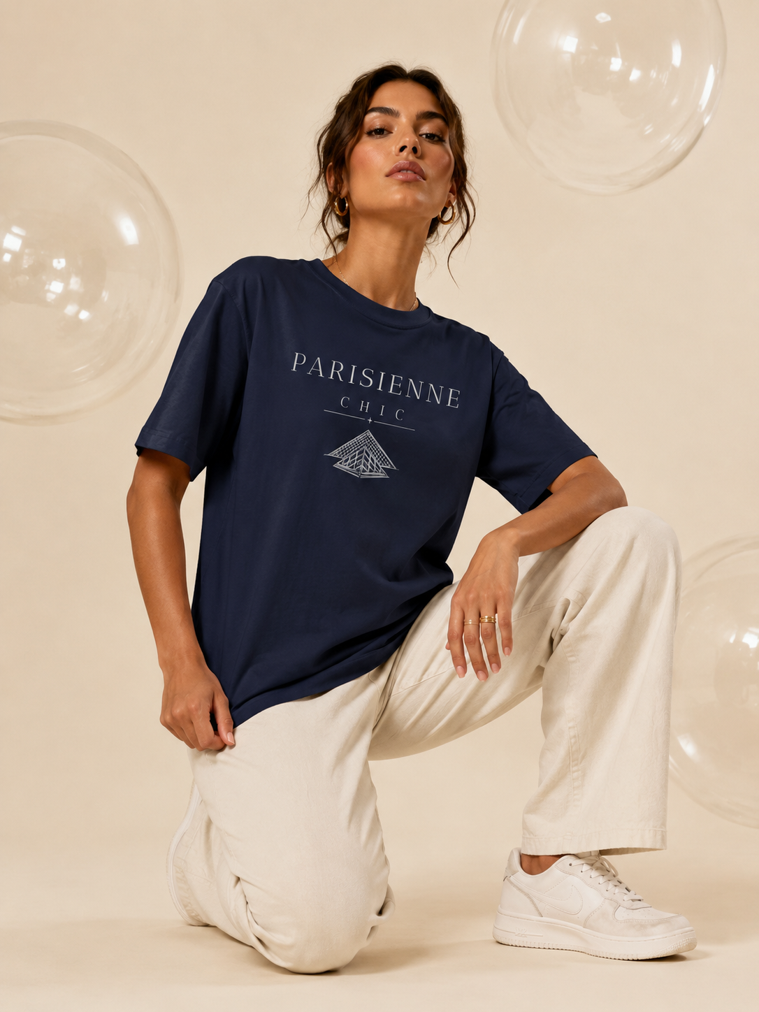 A woman kneeling in a studio wearing a navy blue PARISIENNE CHIC graphic T-shirt with a minimalist pyramid design, paired with cream trousers and white sneakers against a soft beige background with clear decorative bubbles.