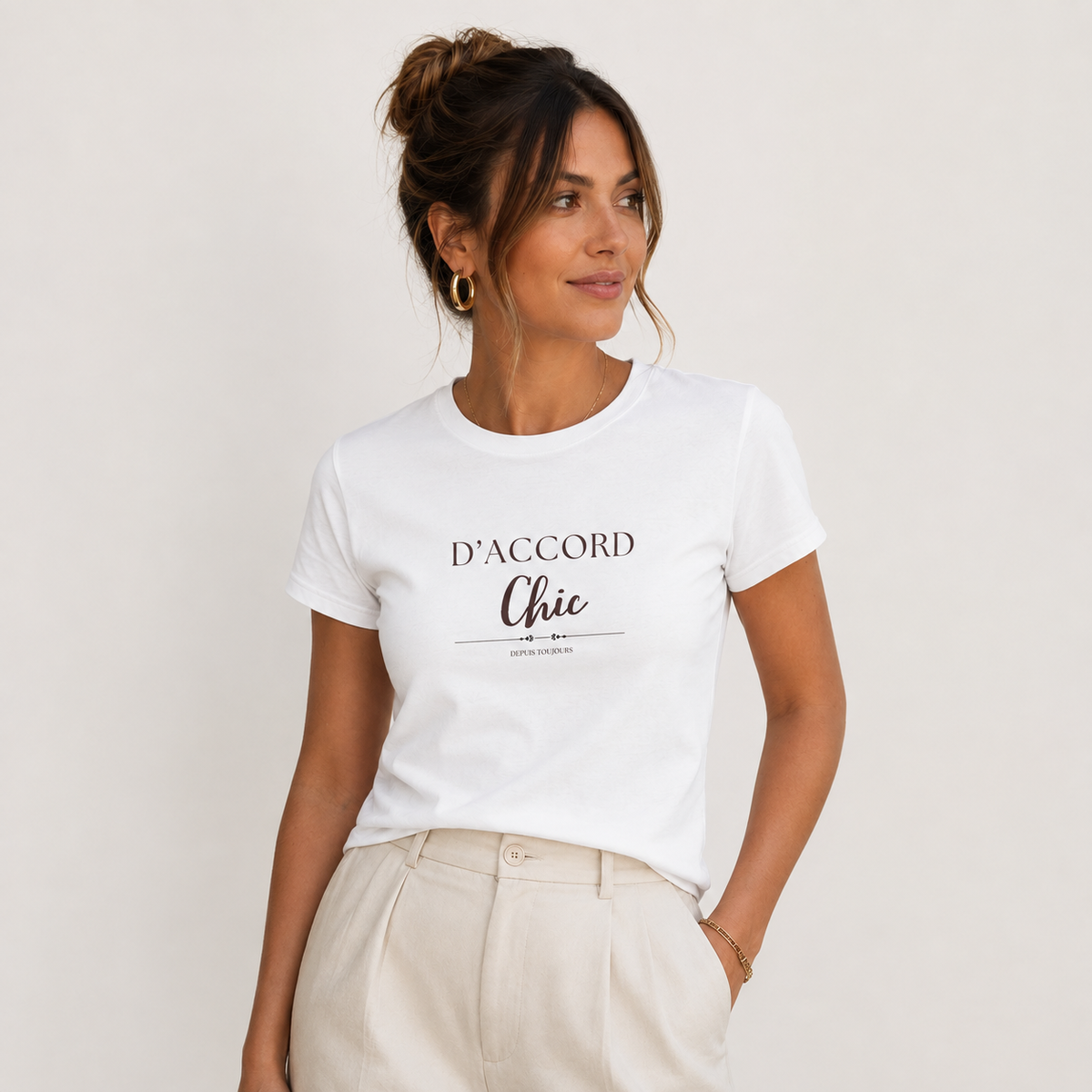 Woman wearing a white D’Accord Chic fitted T-shirt with dark front typography, paired with light beige trousers in a minimalist light gray studio.