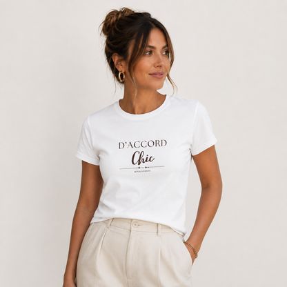Woman wearing a white D’Accord Chic fitted T-shirt with dark front typography, paired with light beige trousers in a minimalist light gray studio.