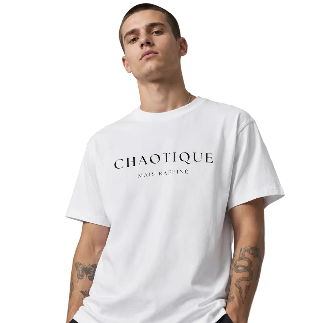 Man wearing a white crewneck T-shirt with black “CHAOTIQUE MAIS RAFFINÉE” slogan print, styled with black pants against a neutral gray studio background.