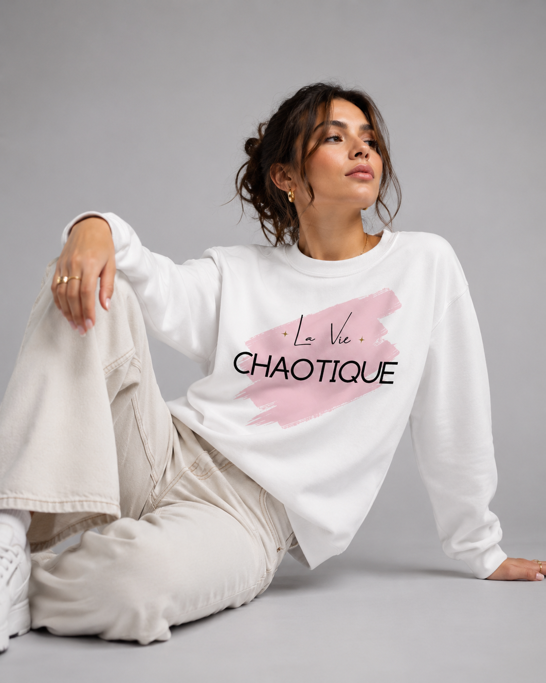 A woman sitting on a gray studio floor wearing a white crewneck sweatshirt with a pink brushstroke graphic and the text 'La Vie CHAOTIQUE' in black, paired with beige wide-leg sweatpants.